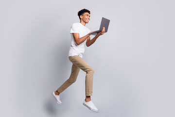 Full size profile photo of brunet optimistic curly guy jump hold laptop wear white t-shirt pants isolated on grey background