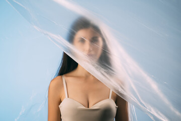 Skin rejuvenation. Plastic surgery. Body enhancement. Art portrait of woman cleavage in beige lingerie face hidden behind wrinkled texture polyethylene film isolated on blue background. © golubovy
