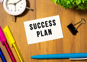 A business card with the text SUCCESS PLAN lies on a wooden office table among office supplies.