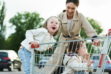 Happy mother and her daughters are having fun with a shopping cart on a parking lot beside a...