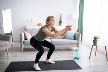 Positive senior lady doing squats on domestic workout in living room, free space