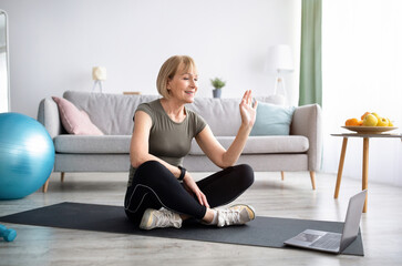 Mature woman in sports clothes sitting on yoga mat in front of laptop, ready for domestic workout, greeting her trainer