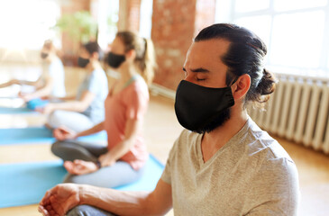 fitness, yoga and health care concept - man with group of people wearing black face protective masks for protection from virus disease meditating in lotus pose at studio