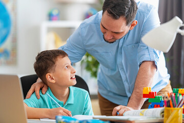 happy father helps his son with his homework using a laptop during distance learning. A parent explains a new topic to a child in an online lesson. Achieving excellence together and enjoying success