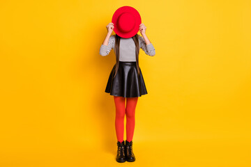 Full length photo of funny childish young girl dressed leather skirt hiding red headwear isolated yellow color background