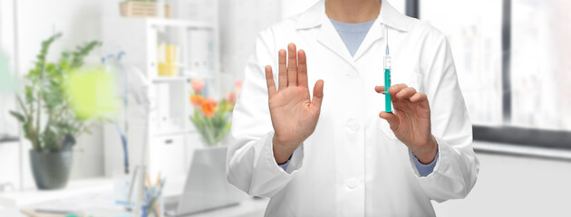 medicine, vaccination and healthcare concept - close up of female doctor or nurse with syringe showing stop gesture over medical office at hospital background