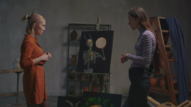 Professional female art critic viewing contemporary artwork of lovely woman painter, analyzing, commenting paintings and giving recommendations due to her viewpoint in art workshop.