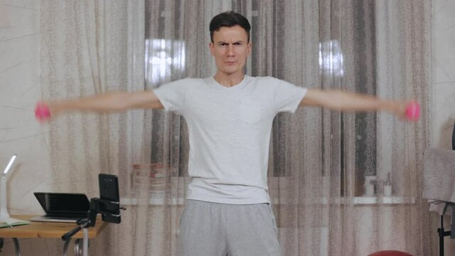 Serious slim male athlete in sportswear standing at home and doing lateral raise exercise with dumbbells while looking at camera 