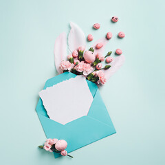 Easter card note with blue envelope, bunny ears and fresh flowers on pastel background. Creative Easter concept. Minimal message with copy space for text. Flat lay.