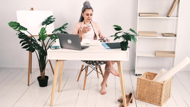Attractive young barefoot caucasian woman in a pink jacket sits in an office at a table with an open laptop. She answers the phone putting her feet on the table. Slow motion, 4k