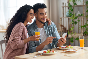 Fototapeta na wymiar Cheerful Arab Spouses Using Smartphone While Eating Breakfast In Kitchen