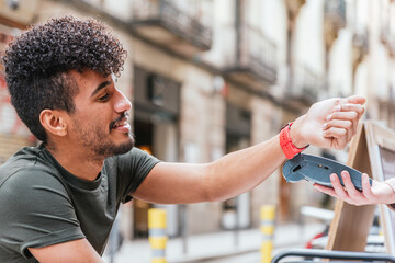 attractive young latin man using a watch to pay. He is in the terrace of a cafe at the street.