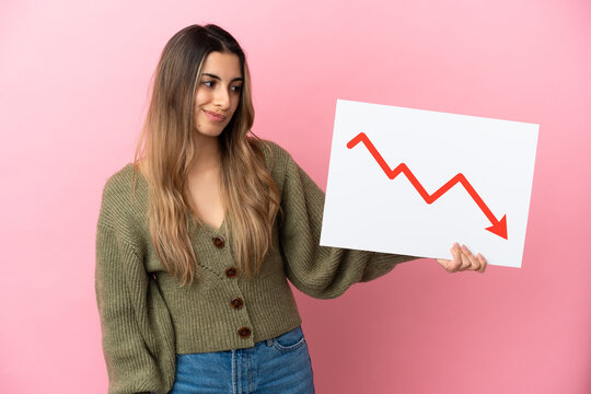 Young caucasian woman isolated on pink background holding a sign with a decreasing statistics arrow symbol with sad expression