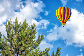 A hot air balloon over green forest on background of fluffy white clouds and blue sky.