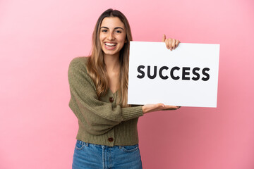 Young caucasian woman isolated on pink background holding a placard with text SUCCESS with happy expression