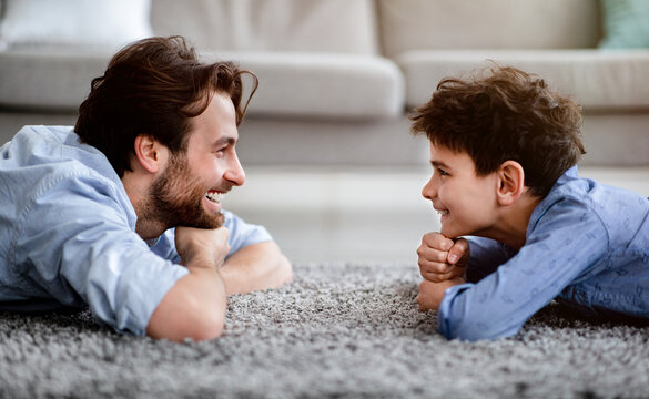 Two generations. Profile portrait of happy father and his son lying on carpet at home and looking at each other, side view