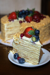 The piece of multi-layered cake decorated with mixed berries. Blueberries, strawberries, redcurrant on small plate on dark brown background. Soft selective focus, copy space.