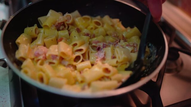 Man holding black plastic spoon and stirring tasty carbonara pasta in cooking pan, traditional italian recipe of mezze maniche pasta with eggs sauce and frying bacon. Italian pasta preparation