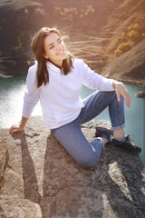 Attractive cute young caucasian woman sitting on stones in the mountains against the background of a high mountain lake on a sunny day