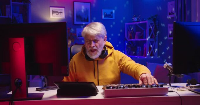 Portrait of cheerful Caucasian senior man DJ enjoying hobby playing music at home recording studio workshop creating beat sound in neon light using professional mixer console equipment on vinyl plate