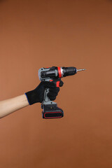 Female hand in a working rag glove holds an electric screwdriver on a battery on a brown background