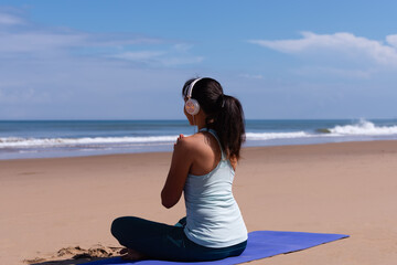 people, fitness, music and healthy lifestyle concept - asian girl meditating listening the music with headset on tropical beach and blue sky background.