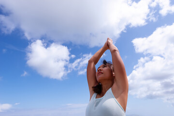 people, fitness, sport and healthy lifestyle concept - asian girl making yoga pose with arms up over blue sky background