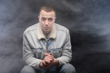 young man sits on a chair covered in smoke in the studio on a black background
