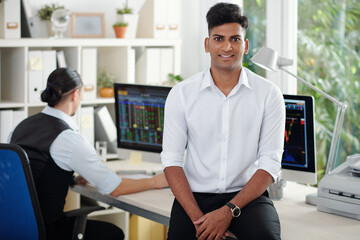 Fototapeta premium Portrait of happy handsome young Indian stock broker leaning on office table and smiling at camera