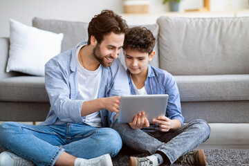 Father and son using tablet computer at home, checking new application, sitting on floor carpet...
