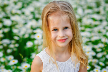 Fototapeta na wymiar Portrait of a beautiful little girl with blonde hair and blue eyes in a white summer dress on a field. A child in a flowery meadow. Copy space.