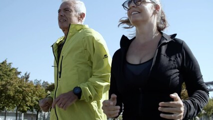 Couple of happy older runners jogging down street. Senior people in sporty clothes running in morning together. Dolly shot. Elderly age and lifestyle concept - Powered by Adobe