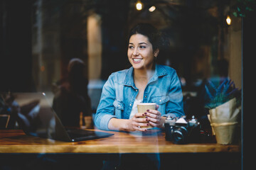 Cheerful hipster girl with takeaway coffee sitting at cafeteria table with modern laptop and camera technology and smiling, window view of funny female freelancer enjoying leisure for working remotely