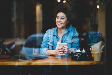 Joyful hipster girl with takeaway coffee sitting at cafeteria table with modern laptop and camera technology and smiling, window view of happy female freelancer enjoying leisure for working remotely
