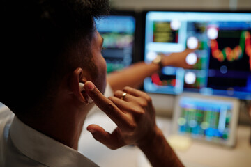 Trader with earbuds pointing at computer screen with stock data and buying assents