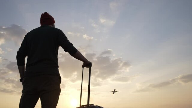 Man with a suitcase at the airport at sunset, sunrise. A man stands and watches as a large passenger plane takes off. Landscape with a beautiful sky with clouds on which the plane flies and a man stan