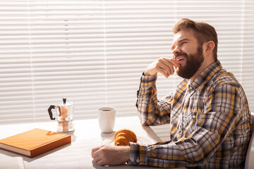 Side view of pensive young bearded male businessman drinking cup of coffee on background of blinds. Concept of pleasant morning or lunch break.