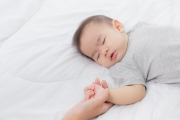 Young asian mother touch hand of little baby girl with tender on bed in the bedroom, mom love newborn and care, woman with expression with child together, parent and daughter, family concept.