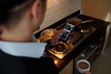 Man drinking morning coffee in dark kitchen and checking stock market updates via application on smartphone