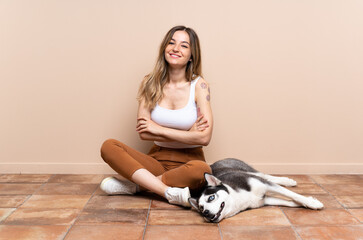 Young pretty woman with her husky dog sitting in the floor at indoors keeping the arms crossed in...