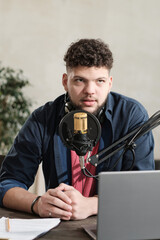 Young male radio dj sitting at the table with laptop and speaking in microphone during his work
