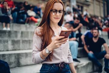 Obraz premium Puzzled female blogger in optical eyewear checking received email message with travel checklist using smartphone device with 4g internet connection, confused hipster girl making mobile booking