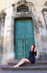 Fototapeta na wymiar Redhead European girl sits at the ancient green door in the old European town in summer