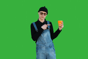 Happy young man in casual clothes on a background of green wall shows thumb up and holds eco cup and smiles while looking at the camera