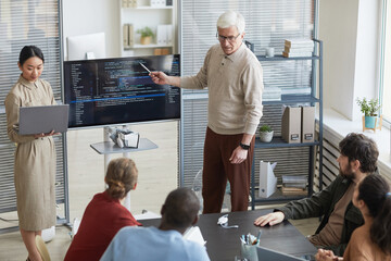Portrait of modern senior man giving presentation in office to diverse IT team and pointing at code...