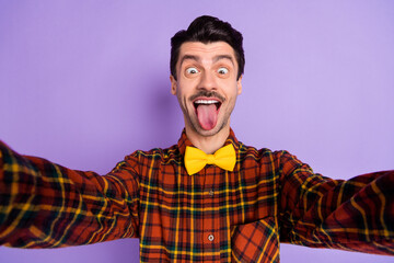 Photo of funny excited guy take selfie tongue out mouth wear bow tie checkered shirt isolated violet background