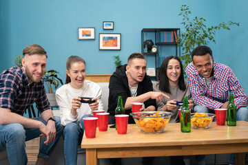 A multinational group of college students sit on the couch and play video games on the TV at home. Smiling friends hang out. The girls compete in a video game.