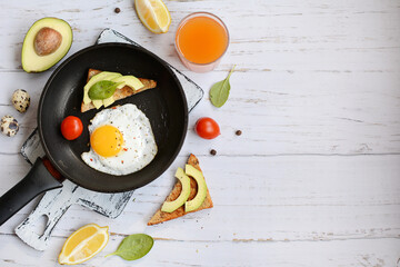 Composition of a frying pan with scrambled eggs, toast with avocado and tomatoes on a light wooden background with copy space. View from above