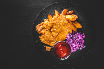 Homemade breaded viennese schnitzel with potato and salad on a plate on black wooden table...