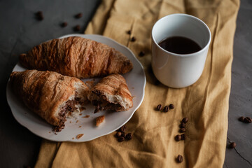 white cup of coffee, two croissants on a white plate. broken croissant with chocolate. breakfast on a textured gray surface with a yellow cloth. Morning meal and breakfast concept